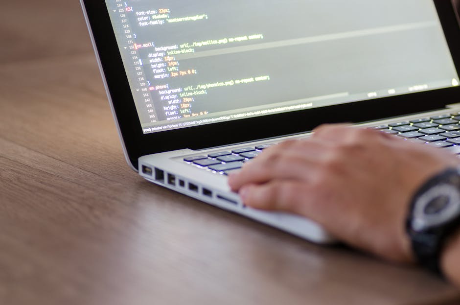 A close-up view of a person's hand wearing a black wristwatch, resting on a laptop keyboard with a visible screen displaying lines of code or programming language. The laptop is placed on a wooden sur